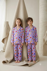 Twin girl and boy wearing matching lavender purple linen button-down pajama sets featuring a vibrant garden fruit print with white piping.