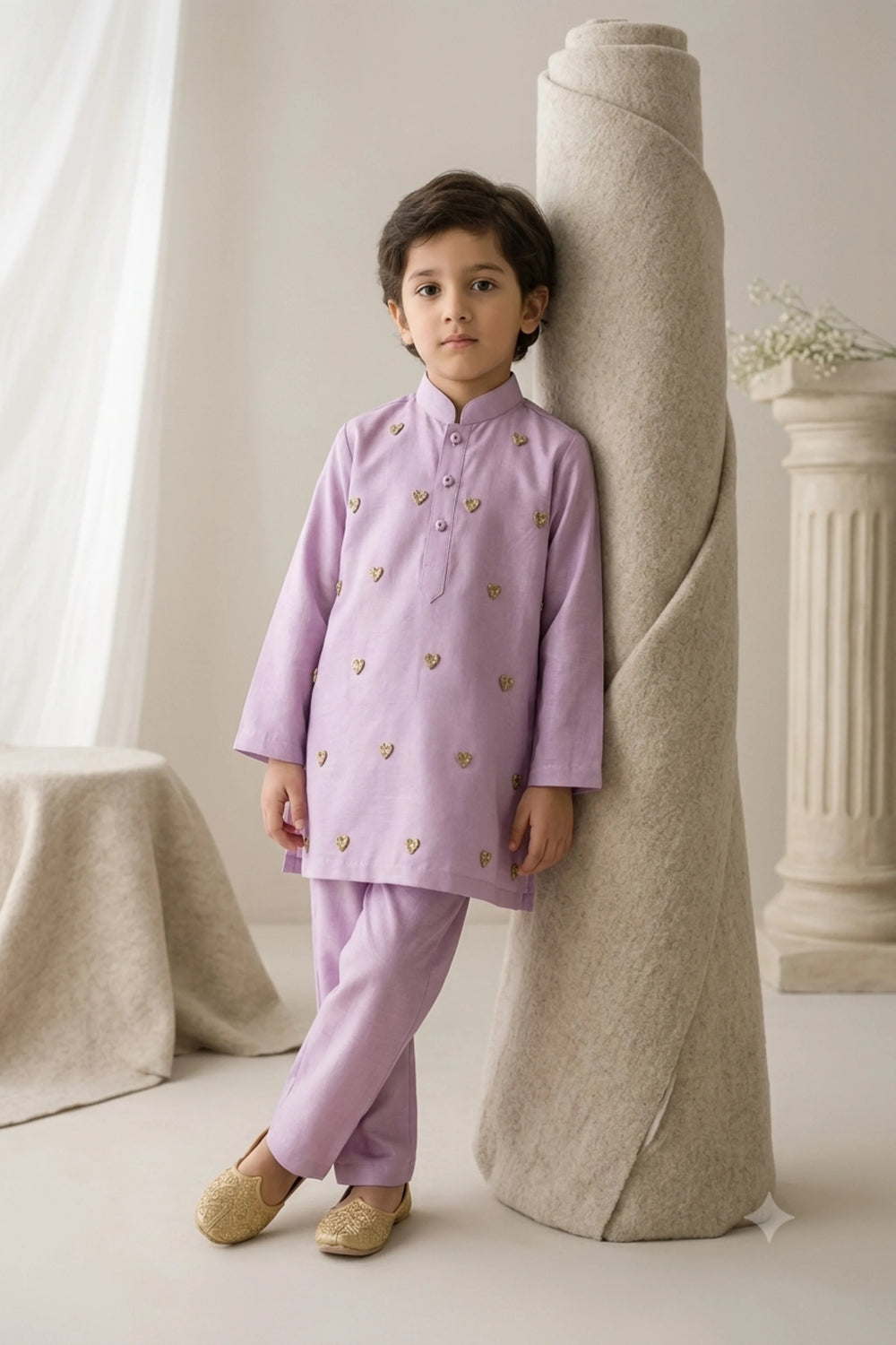 Young boy leaning against a neutral roll wearing a lilac Tussar silk kurta pajama set embellished with delicate golden heart motifs paired with gold khussas.
