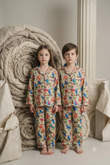 Twin girl and boy wearing matching beige linen button-down pajama sets featuring a colorful botanical bird print with contrast piping.