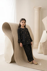 Young girl standing leaning on a textured roll wearing a midnight black shamoz silk kurti and trouser set embellished with golden teardrop booti motifs and gold khussas.