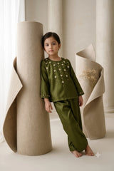 Young girl leaning against a cylindrical roll wearing a deep olive green shamoz silk kurti and trouser set embellished with geometric crystal and golden motifs.