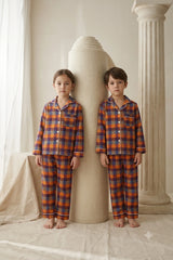 Twin girl and boy standing side by side wearing matching orange and navy blue plaid cotton button-down pajama sets.
